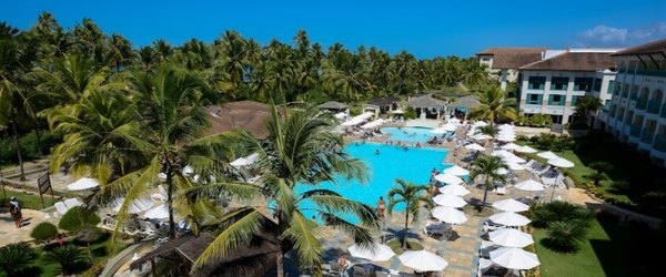 Vista parcial do Costa do Sauípe, com uma de suas piscinas, parte de uma edificação, os típicos coqueiros e o lindo céu azul
